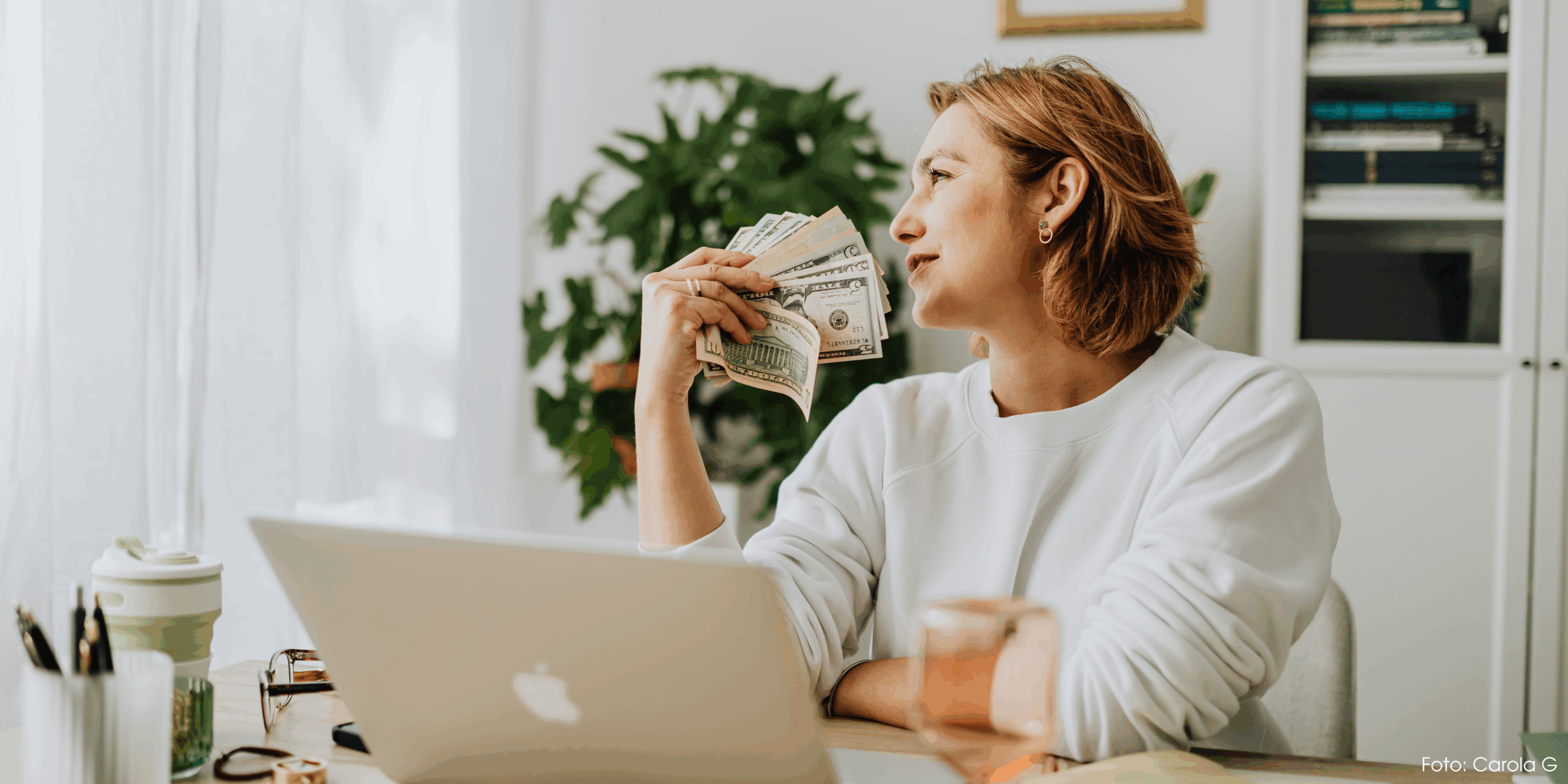 Eine blonde Frau mit kurzen Haaren, in einem hellen Raum am Schreibtisch vor dem Laptop mit einem Fächer aus Geld vor dem Gesicht